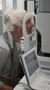 Vertical shot of elderly woman during eye exam in ophthalmologist office at clinic putting head into frame of diagnostic machine