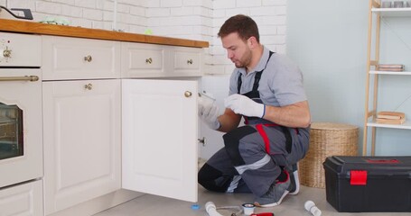 Professional plumber works under the kitchen sink at home, repairing leaking pipes. The maintenance service focuses on plumbing expertise, ensuring functionality and efficient water system repairs.