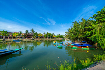 Wooden boats on the Thu Bon river in Hoi An ancient town. Yellow old houses on waterfront reflected in river. Hoi An is a UNESCO world heritage site in Vietnam