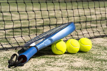 Blue padel racket with three yellow tennis balls by net on court; sunny daylight and selective focus celebrate padel, tennis, sportlife and outdoor lifestyle activity.