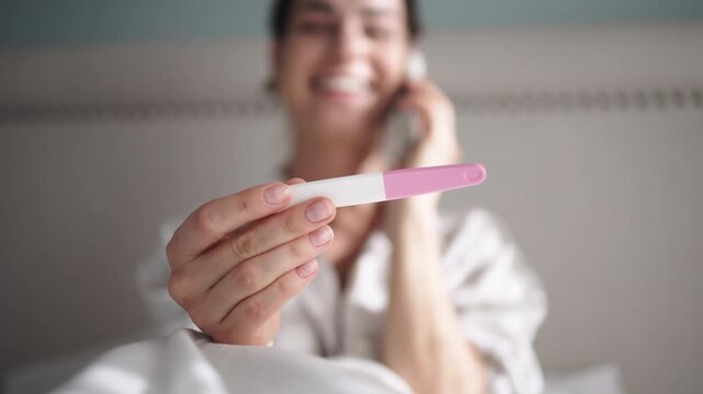 Woman holding pregnancy test with positive result, smiling while sitting on bed, surrounded by soft bedding, conveying joy and anticipation of new beginnings