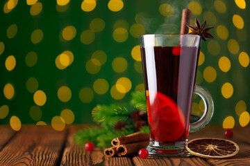 Steaming hot mulled wine with cinnamon stick and slice orange in glass mug against green background of glowing garland lights
