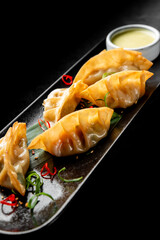 Golden pan-fried Asian dumplings (gyoza, jiaozi, potstickers) served on a black plate with a white dipping sauce, garnished with chili and green onions, against a dark background.