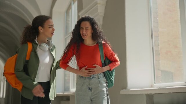 Two Hispanic Latina female students walking university girl holding stomach pain women friends in college talking feeling ache menstrual period painful cramp abdominal health problem medicine gastric