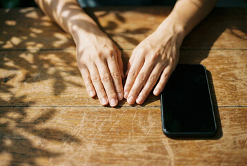 Hands resting on wooden table with smartphone face down relaxing in sunlight