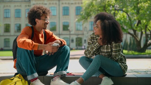 Two happy African American friendly students conversation sitting outdoor multiracial friends African American woman girl female with boy man guy in city talking smiling university college high school