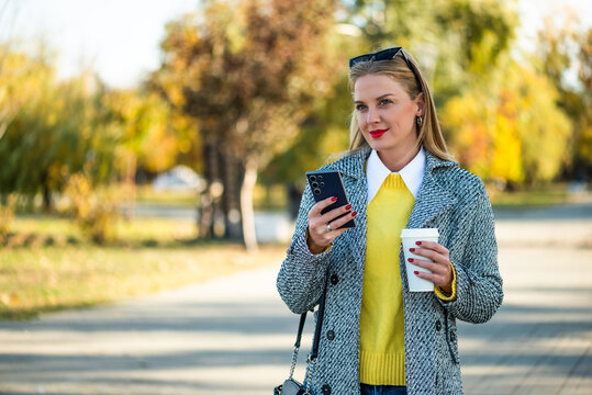 Portrait of modern businesswoman with sunglasses in modern autumn coat using mobile phone and drinking coffee while standing in the city park during sunny day.	