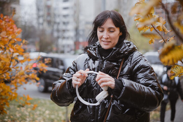 Woman in warm black puffer jacket holds headphones while walking through city park with golden autumn trees, enjoying music outdoors