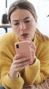 Confused young woman looking at smartphone screen, touching her lip and thinking about a problem or difficult decision while lying on sofa. Vertical video.