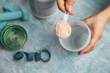 Fototapeta premium Female hand putting scoop of chocolate whey protein powder into shaker on blue background with protein jar, dumbbells and measuring tape, concept of sport nutrition