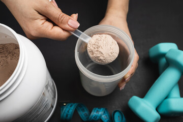 Female hand putting scoop of chocolate whey protein powder into shaker on dark background with...