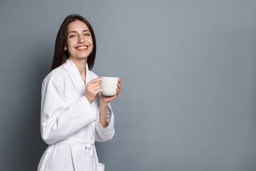 Woman in bathrobe with cup of coffee on grey background. Space for text