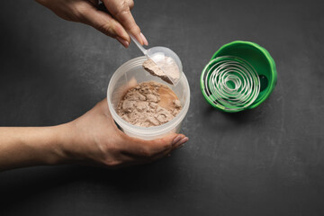 Preparing protein milkshake, female hand putting scoop of chocolate whey protein powder into shaker with milk on dark background, concept of sport nutrition