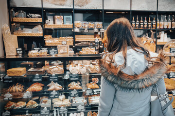 A woman stands in a bakery examining a variety of sweet cakes, croissants, and buns. She is deciding on a dessert to purchase for dinner.