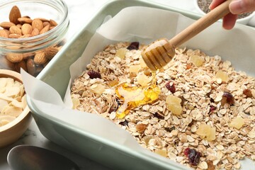 Making granola. Woman pouring honey onto oat flakes and dried fruits at white table, closeup