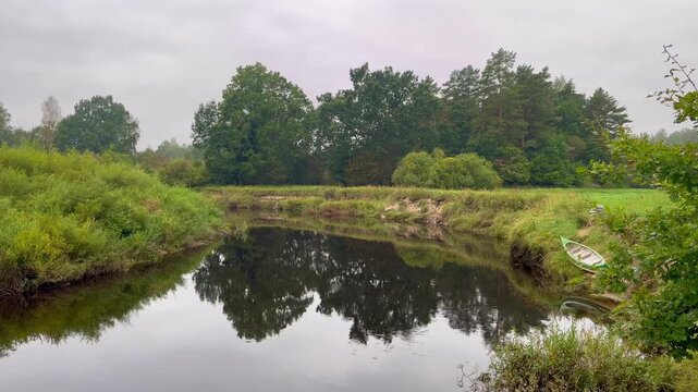 Calm Irbe River in Latvia curving through dense greenery with lone green kayak pulled onto grassy bank, dark reflective water and soft overcast atmosphere in quiet wilderness
