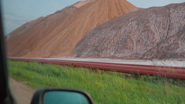 Driving shot revealing huge artificial mountains of salt, a byproduct of potash fertilizer production, towering over the landscape next to an empty road under a cloudy evening sky