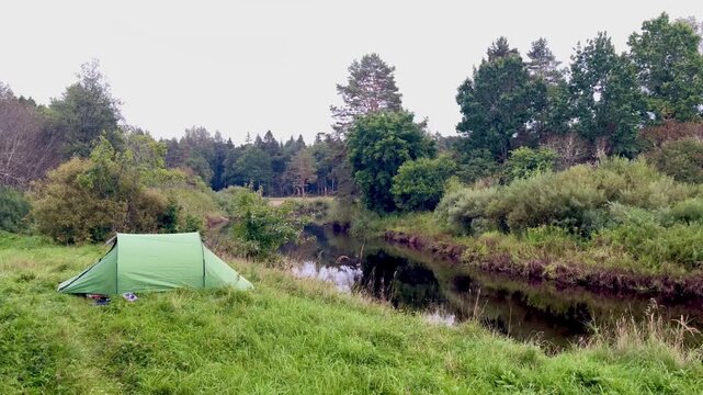 Green tent set on grassy riverbank above calm Irbe River in Latvia, surrounded by dense forest, soft morning light and reflective water creating peaceful wild-camp atmosphere