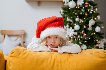 Home portrait of a child girl in a Santa hat. The concept of New Year's holidays. A child peeks out from behind the sofa against the background of a Christmas tree. Christmas mood.
