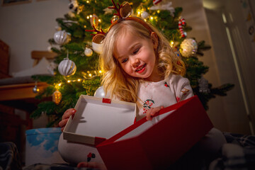 Cute Caucasian child little girl in the dark by a Christmas tree with a garland opens a gift box and rejoices