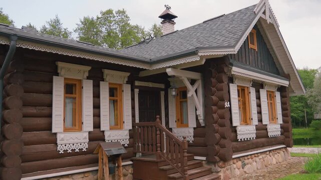 Beautifully crafted traditional russian log house featuring intricate white carved wooden window frames and shutters, set in a rustic countryside environment with surrounding green trees