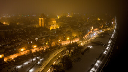 Widok z drona na nocne Stare Miasto w Toruniu, spowite delikatną mgłą. Podświetlone budynki i zabytkowa architektura lśnią w mgiełce, tworząc tajemniczy i nastrojowy miejski pejzaż.