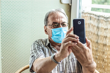 Senior man wearing protection face mask while relaxing at home in the living room and taking a selfie with smart phone.