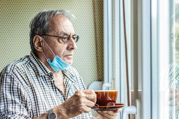 Senior male looking out of the window with a face mask on and a cup of fresh coffee.