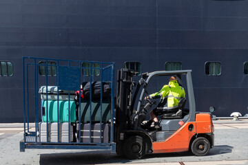 A forklift driver or handler on the quay with moored cruiseship as backdrop handling a forklift cage filled with suitcases of passengers for the ship
