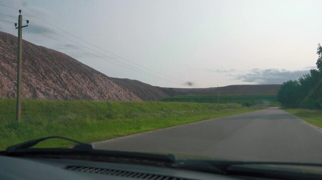 Personal perspective driving along a quiet rural road, passing a large industrial slag heap from a mining operation under a bright sky, illustrating travel through a modified landscape