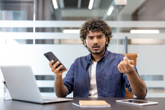 Portrait of a young worried man sitting at a desk in the office, holding a phone in his hand and waving his arms at the camera in frustration