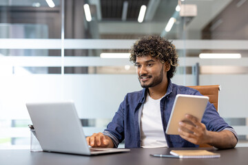 Smiling young Muslim man working in the office at a laptop and using a tablet