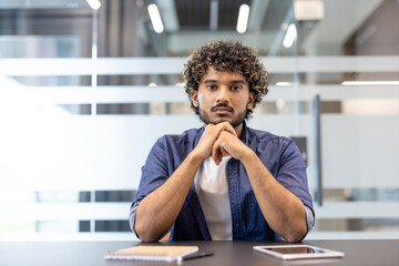 Portrait of a serious young Muslim male teacher and business coach sitting in the office at a desk in front of the camera with a notebook and tablet and looking at the camera
