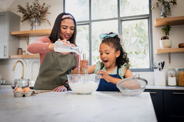 Mother and daughter pouring milk from glass jug into bowl and whisking flour at kitchen island