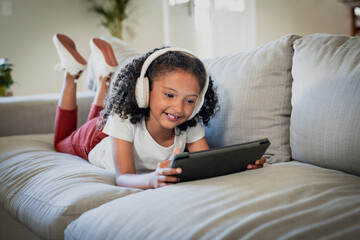 African American girl lying on couch in living room using tablet wearing headphones near plant