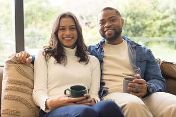 Diverse couple smiling while holding ceramic mugs on upholstered sofa in living room by window