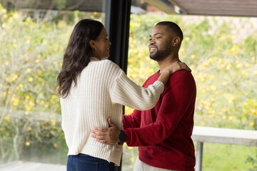 Diverse couple embracing on modern veranda through glass wall by railing overlooking yellow trees