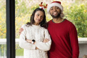 Diverse couple standing near sliding door at home wearing antler headband Santa hat smartwatch