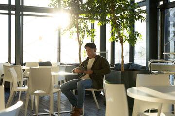Asian man sitting among white chairs in modern lounge holding digital tablet next to potted tree
