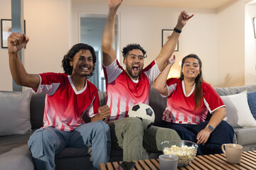 Friends wearing red jerseys sitting at home cheering holding soccer ball popcorn mugs smartphone