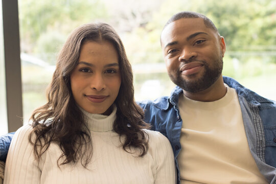 Diverse couple wearing denim jacket sitting on couch at home overlooking garden through window - Powered by Adobe