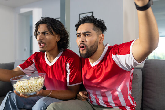 Male friends wearing red jerseys cheering on gray sofa in living room with bowl of popcorn - Powered by Adobe