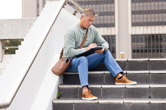 Man sitting on office steps sketching in black portfolio with green pencil and coffee cup