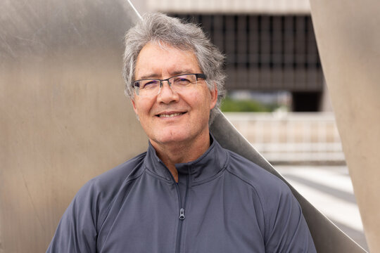 Mature man standing in front of angular metal sculpture at plaza wearing eyeglasses, zip-up jacket