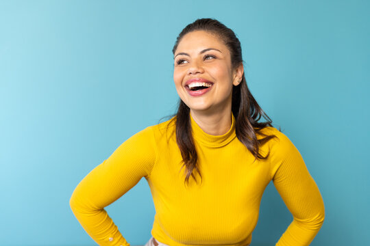 Woman wearing yellow turtleneck sweater, smiling, posing hands on hips in studio with blue backdrop