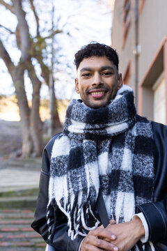 Man in twenties standing on stone steps amid bare trees wearing leather jacket and checkered scarf