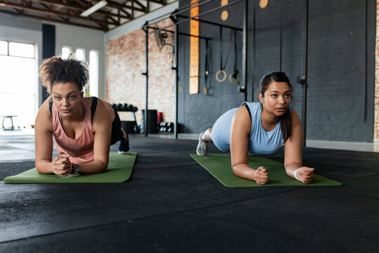 Diverse female friends holding plank on green mats in fitness studio by pull-up rig with dumbbells