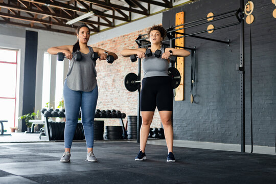 Diverse female friends wearing athletic wear holding dumbbells performing upright row in gym