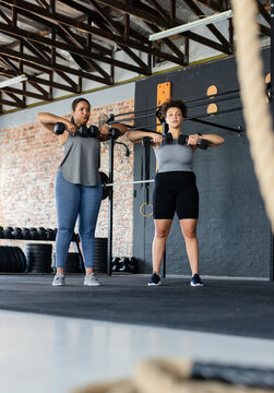 Diverse female workout partners performing upright rows in gym with dumbbells and climbing rope