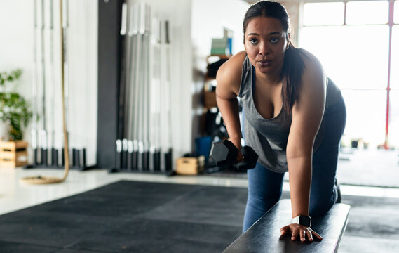 African American woman in sportswear lifting dumbbell, leaning on bench beside weight racks in gym